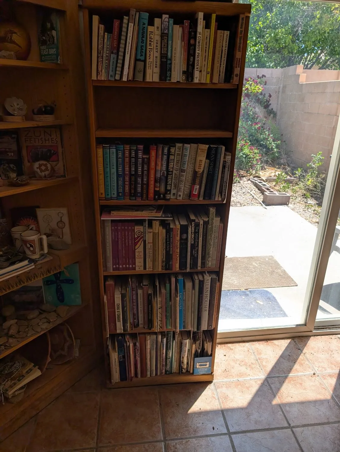 A packed bookcase next to a sliding glass door in a typical Albuquerque home before a free pickup by Josh Eldred and the New Mexico Literacy Project — five shelves of hardcovers, paperbacks, and cookbooks with a sunny courtyard visible through the glass