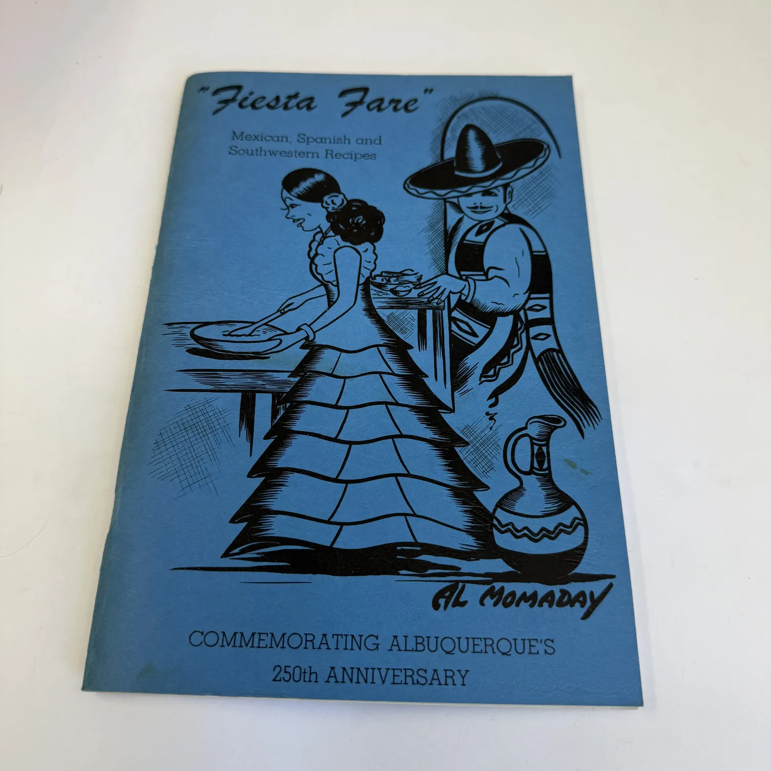 The cover of Fiesta Fare: Mexican, Spanish and Southwestern Recipes, a 1956 Albuquerque 250th-anniversary commemorative cookbook with a black-and-blue illustration of a Mexican-dressed woman cooking and a man in a sombrero, signed Al Momaday in the lower right.