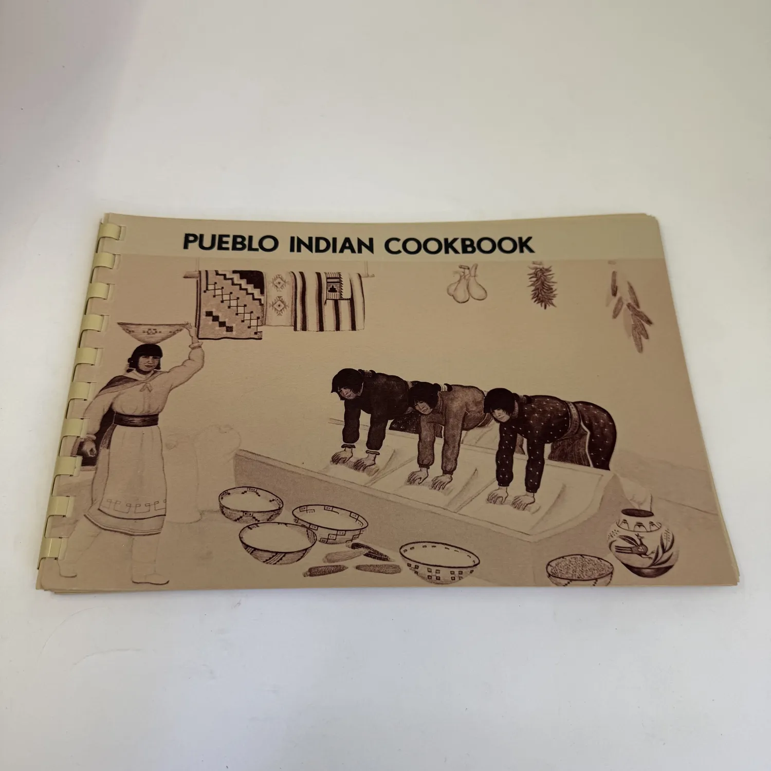 A spiral-bound copy of Pueblo Indian Cookbook by Phyllis Hughes, published by the Museum of New Mexico Press, with a sepia-toned illustration on the cover showing women preparing food with traditional Pueblo pottery and weavings.