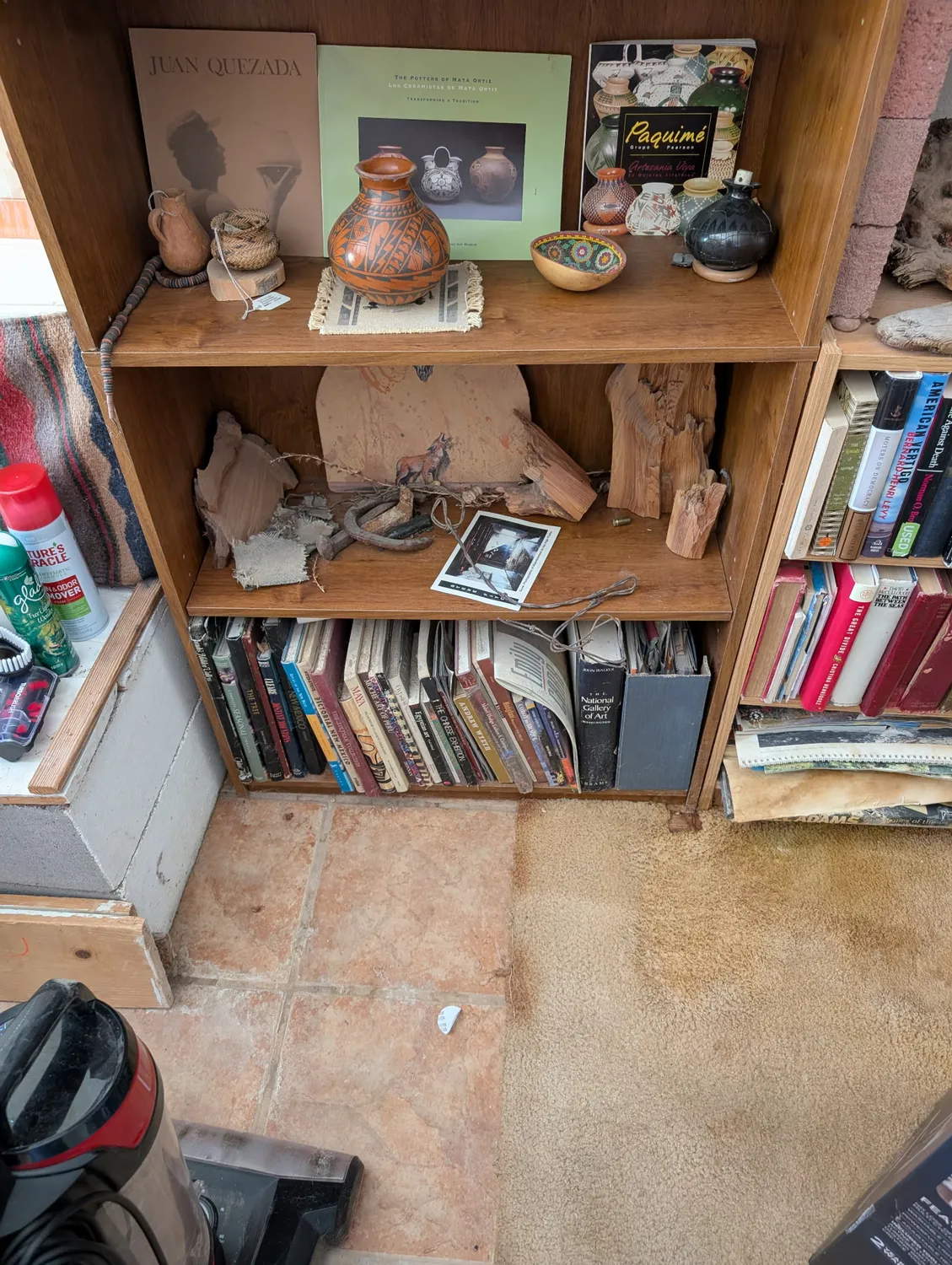 A New Mexico home bookshelf during a senior downsizing evaluation by Josh Eldred of the New Mexico Literacy Project — a Juan Quezada pottery book and Paquimé ceramics displayed on the top shelf alongside handmade Mata Ortiz pots, natural artifacts and a framed photograph on the middle shelf, and well-read books below on saltillo tile flooring