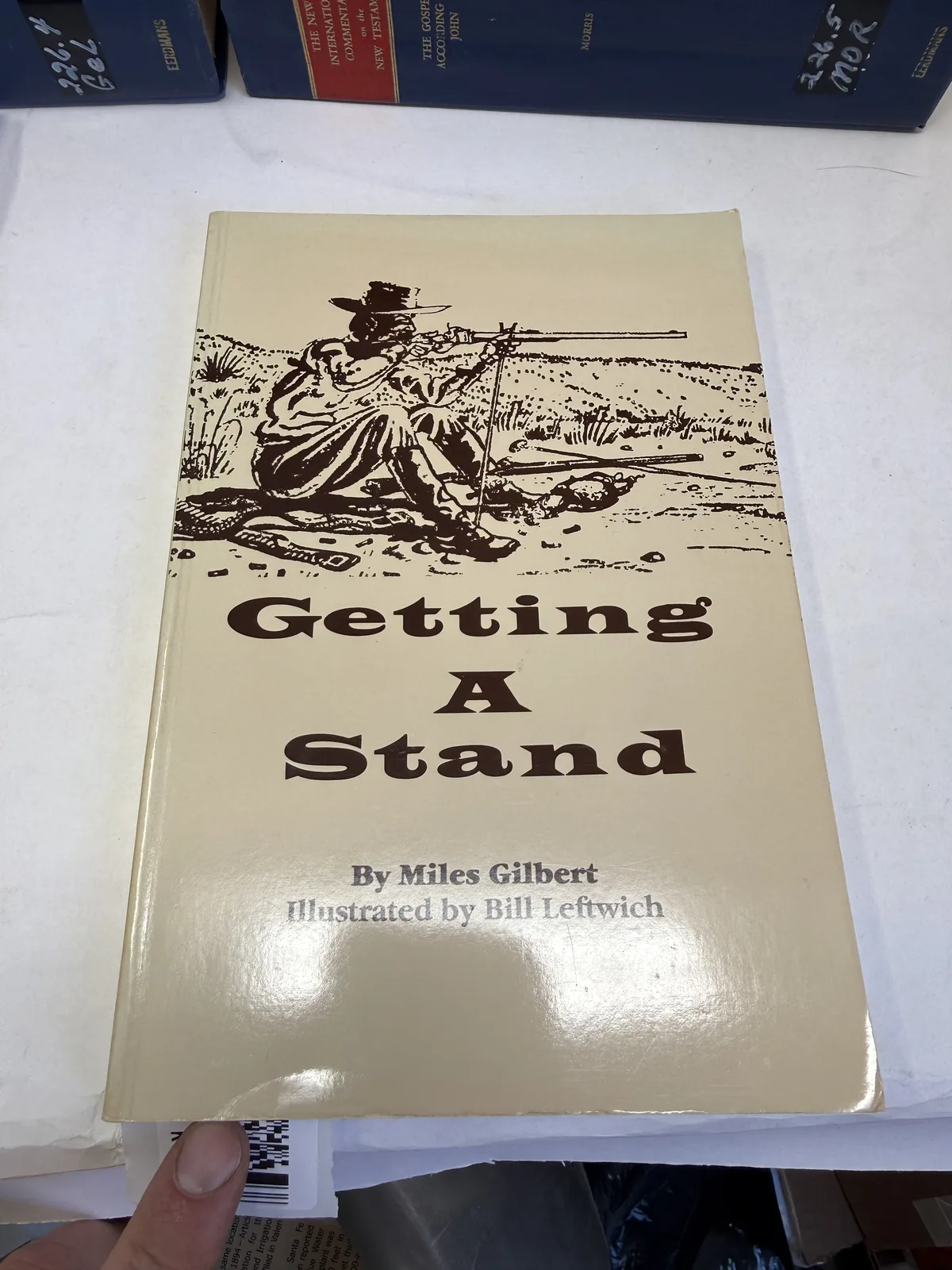 Cover of Getting A Stand by Miles Gilbert, illustrated by Bill Leftwich, with a drawing of a buffalo hunter firing from a sitting rest, photographed at New Mexico Literacy Project