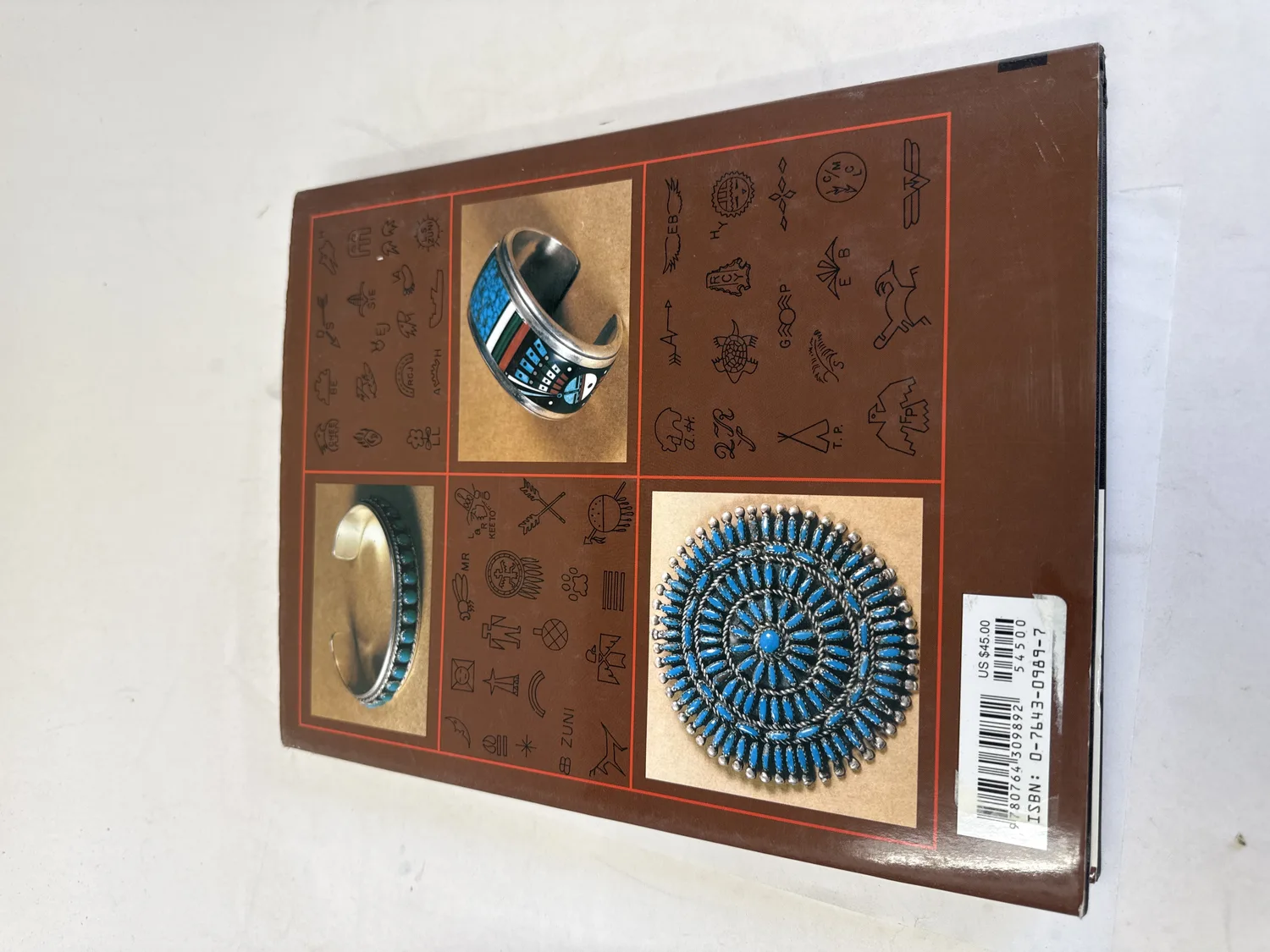 Back cover of Hallmarks of the Southwest showing sample hallmark symbols surrounding photographs of turquoise and silver jewelry including a Zuni petit-point cluster brooch and channel-inlay cuff bracelet, with ISBN barcode 0-7643-0989-7 and US forty-five dollar cover price. Photographed at the New Mexico Literacy Project sorting desk in Albuquerque.