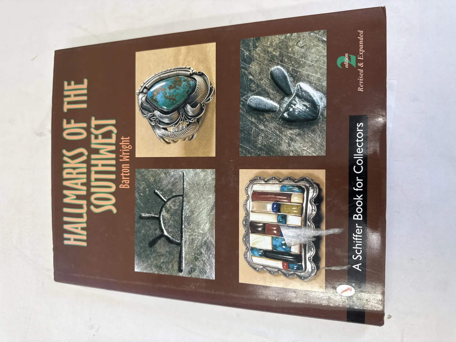 Front cover of Hallmarks of the Southwest by Barton Wright, Revised and Expanded 2nd Edition, showing a turquoise-and-silver bracelet, hallmark stamp dies, and a multi-stone channel inlay belt buckle against a brown background with gold and green typography. Photographed at the New Mexico Literacy Project sorting desk in Albuquerque.