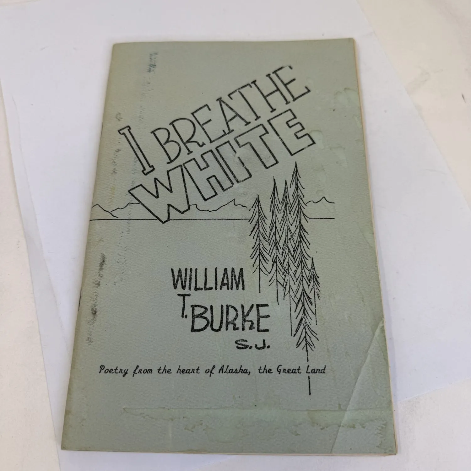 Second angle of the front cover of I Breathe White by William T. Burke S.J., showing the full chapbook with curled lower corner, pale green cover stock with mountain and spruce illustration, Silver Wings Press 1987 Alaska poetry.