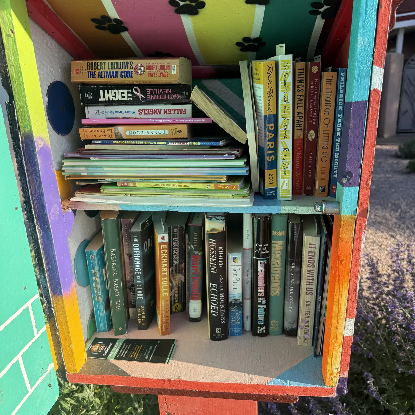 Blue glass-door Little Free Library with two shelves restocked from NMLP intake — Dr. Seuss face-out, vintage Thorndike Desk Dictionary two-volume, NMLP bookmark visible centered