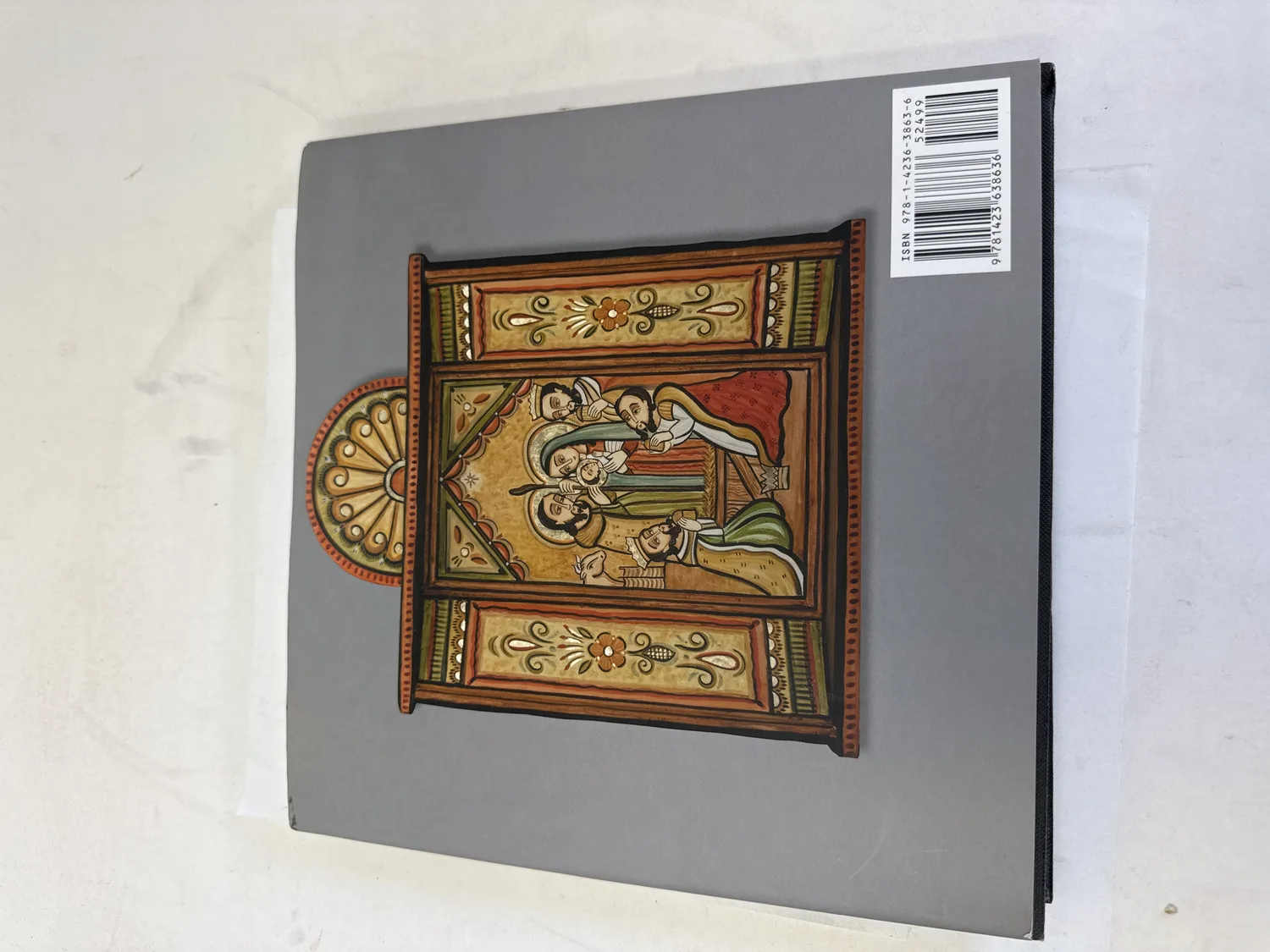 Back cover of Nativities of the Southwest showing a retablo-style painted nativity scene in traditional New Mexico santero style with warm earth tones, arched top, ornamental floral borders, depicting the Holy Family with Magi and shepherds. ISBN barcode 978-1-4236-3863-6 visible. Photographed at the New Mexico Literacy Project sorting desk in Albuquerque.