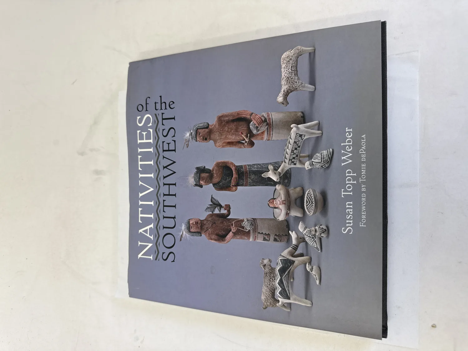 Front cover of Nativities of the Southwest by Susan Topp Weber, showing pueblo-style and ceramic nativity figurines arranged on a blue-gray background, with a stepped zigzag design in the title typography echoing Southwest textile patterns. Gibbs Smith publisher, foreword by Tomie dePaola. Photographed at the New Mexico Literacy Project sorting desk in Albuquerque.