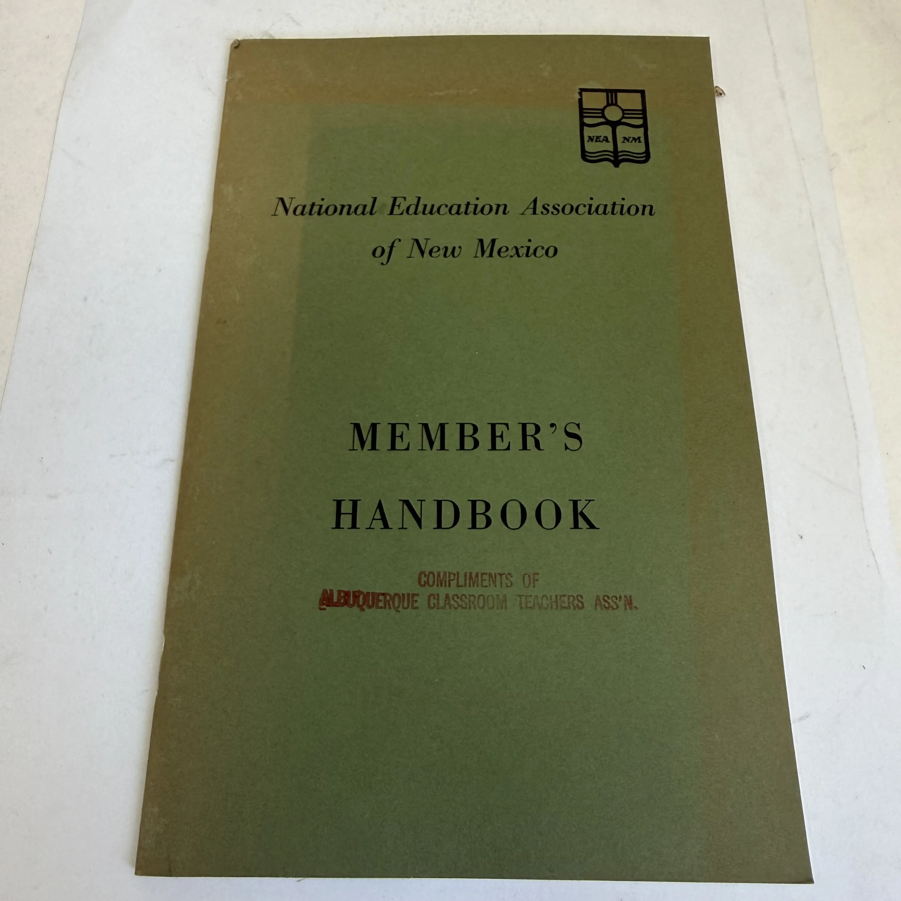 Front cover of the NEA of New Mexico Member's Handbook, olive green staple-bound pamphlet with NEA NM logo showing a book with radiating rays in the upper right. Rubber-stamped in red: Compliments of Albuquerque Classroom Teachers Ass'n. Mid-century institutional ephemera found in a New Mexico estate cleanout by the New Mexico Literacy Project.