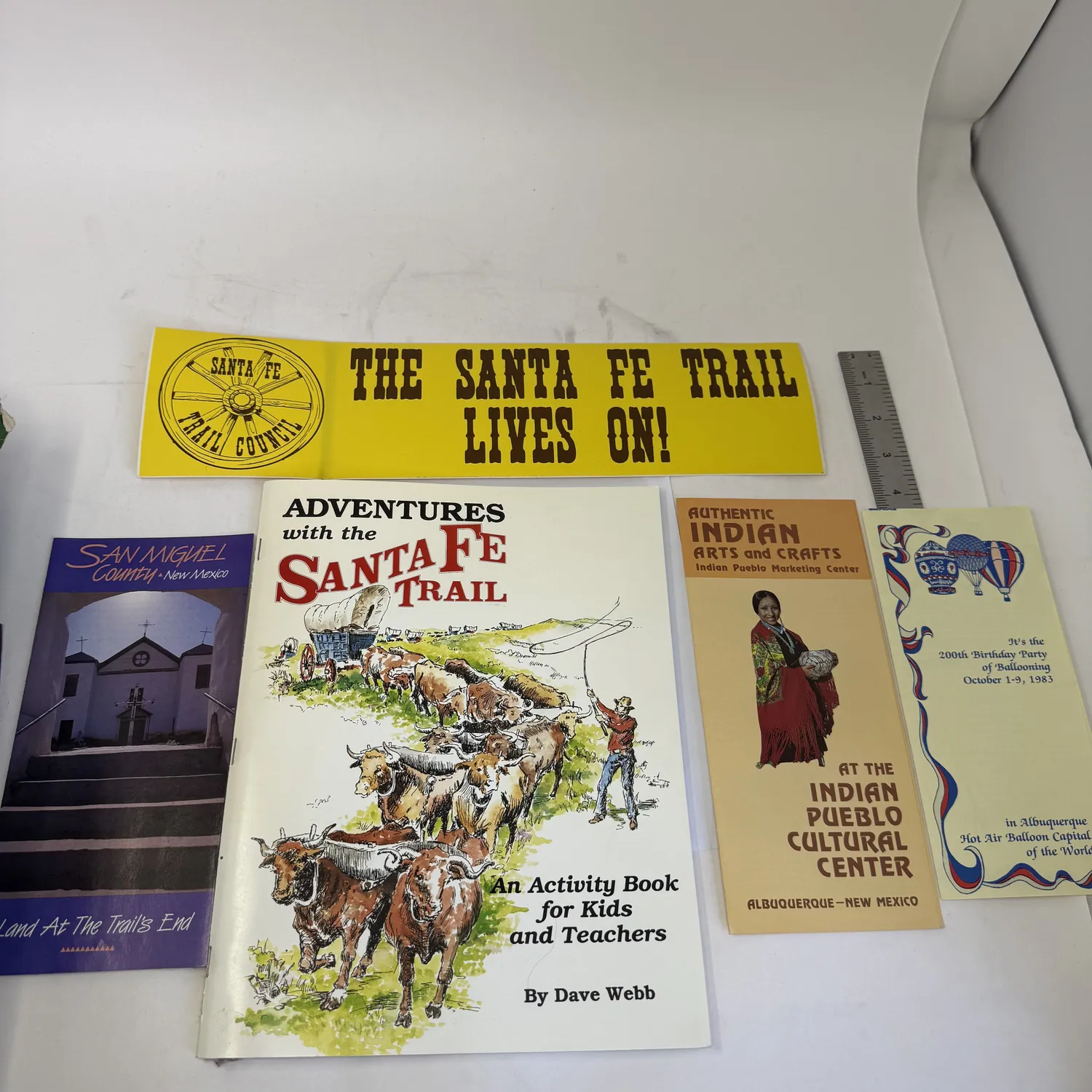 New Mexico ephemera from an estate donation sorted at the New Mexico Literacy Project workspace — Santa Fe Trail Council bumper sticker reading The Santa Fe Trail Lives On, Adventures with the Santa Fe Trail activity book by Dave Webb, San Miguel County New Mexico Land At The Trails End tourism brochure with mission church photograph, Authentic Indian Arts and Crafts at the Indian Pueblo Cultural Center Albuquerque brochure, and 1983 Albuquerque Balloon Fiesta program celebrating the 100th birthday party of ballooning, photographed by Josh Eldred