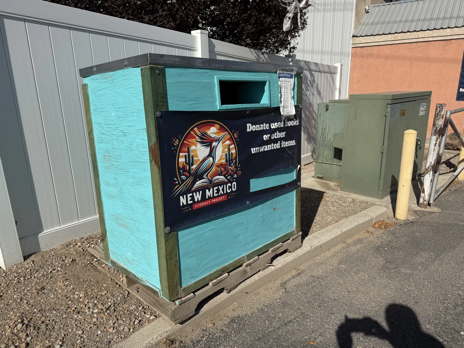 The New Mexico Literacy Project book drop box in Albuquerque, a turquoise wooden box on a pallet with a deposit chute and a roadrunner logo panel reading Donate used books or other unwanted items