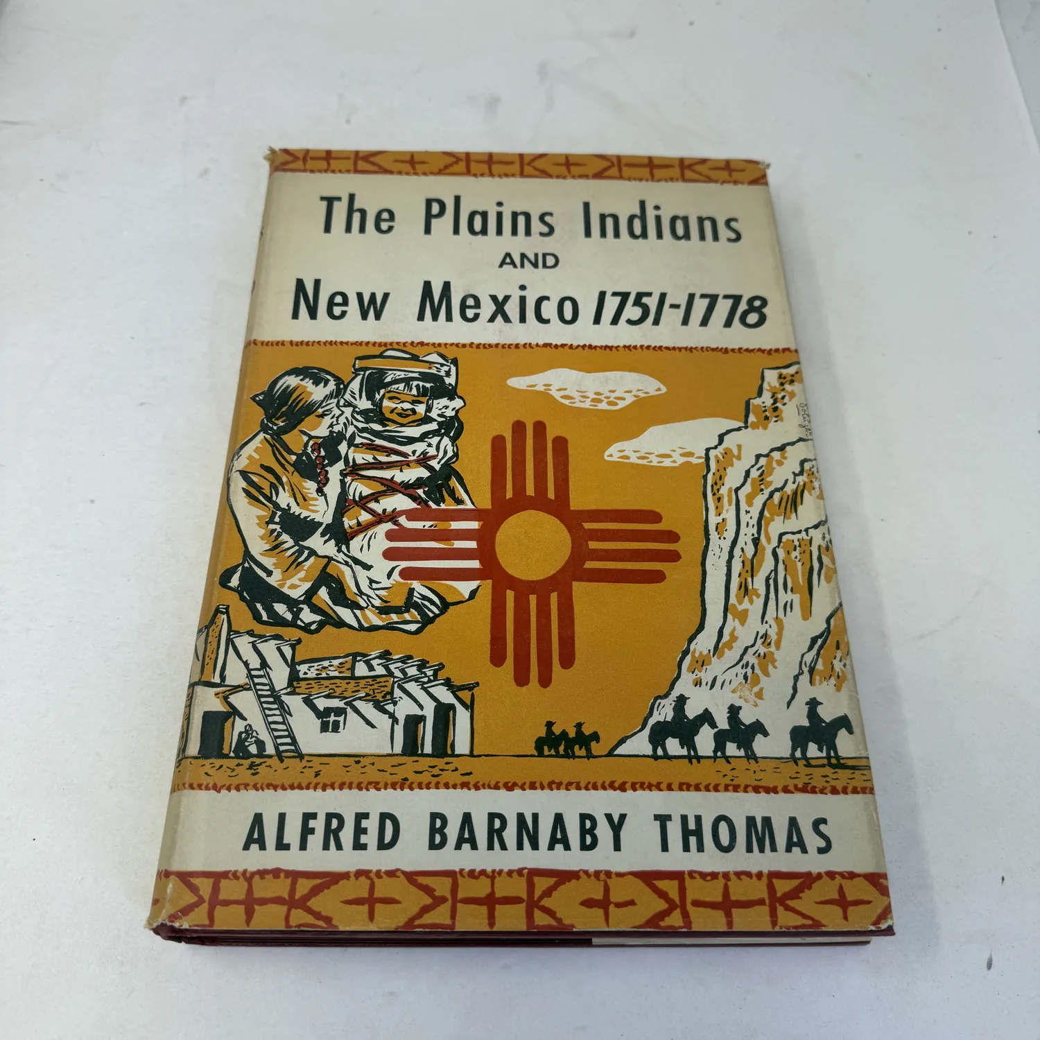 Pictorial dust jacket of The Plains Indians and New Mexico 1751-1778 by Alfred Barnaby Thomas, with a red Zia sun symbol, a Pueblo village and horsemen, photographed at New Mexico Literacy Project
