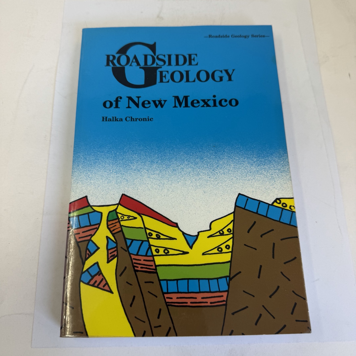 Front cover of Roadside Geology of New Mexico by Halka Chronic — Roadside Geology Series header, colorful stylized geological cross-section cover art showing layered rock formations, fault lines, and volcanic features against blue sky gradient. Mountain Press Publishing Company.