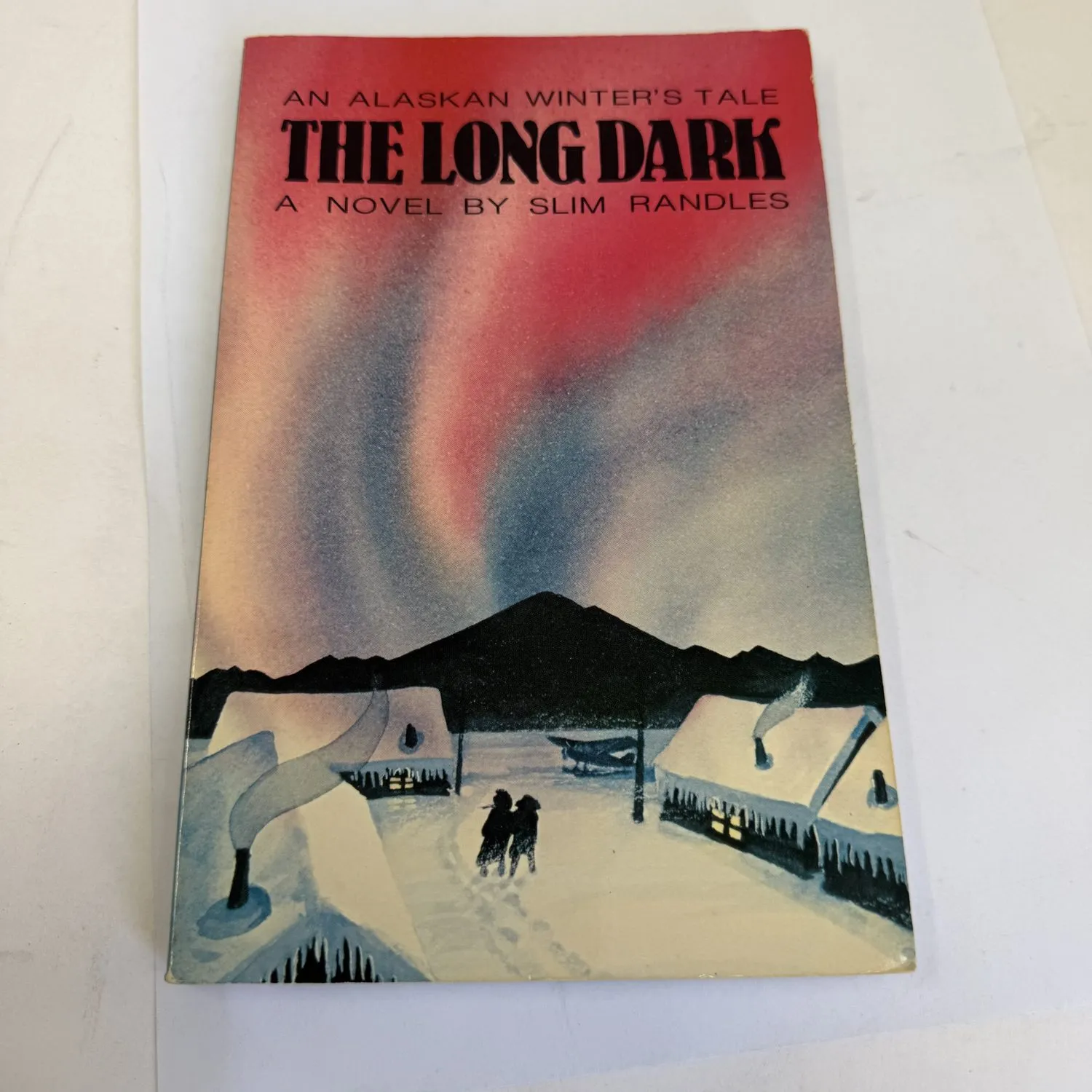 Front cover of The Long Dark An Alaskan Winter's Tale A Novel by Slim Randles, Alaska Northwest Publishing Company 1985, showing a painted aurora borealis scene with red pink and violet Northern Lights streaming over a snowy Alaska Bush village, two silhouetted figures walking between snow-covered cabins toward the light, dark mountain rising behind. Cover art by Sharon Shumacher. Found in a New Mexico estate cleanout by the New Mexico Literacy Project.