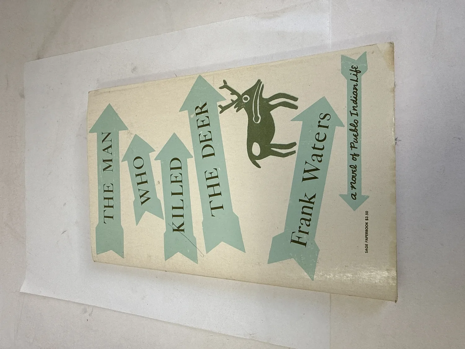 Front cover of The Man Who Killed the Deer by Frank Waters, Sage Paperbook edition circa 1970 — turquoise arrows and stylized Pueblo deer figure on cream background, priced $3.50. Photographed at the New Mexico Literacy Project sorting desk in Albuquerque.