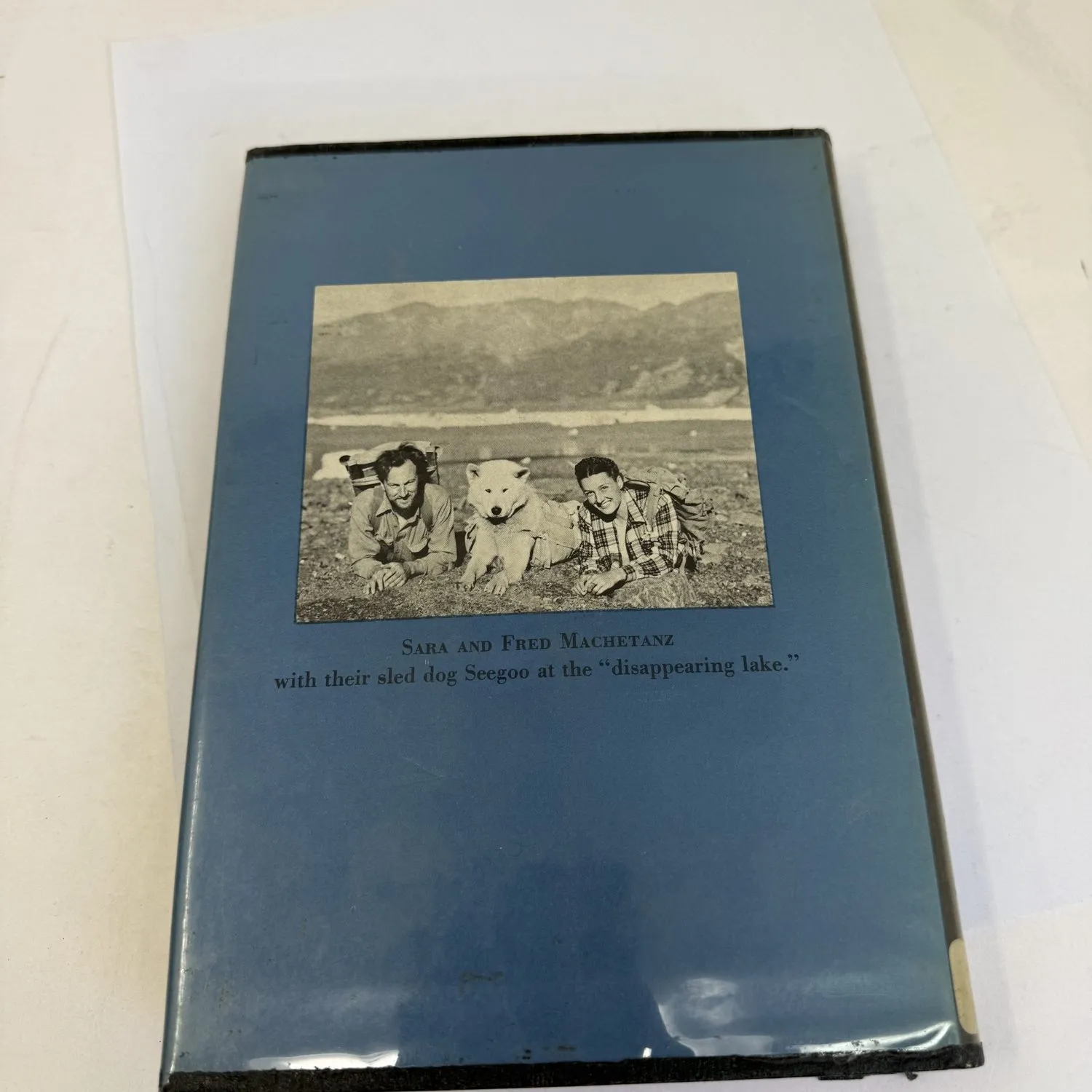 Back of dust jacket of Where Else but Alaska showing Sara and Fred Machetanz lying on the ground with their white sled dog Seegoo at the disappearing lake, mountains in the background. Caption reads Sara and Fred Machetanz with their sled dog Seegoo at the disappearing lake. Charles Scribner's Sons 1954.
