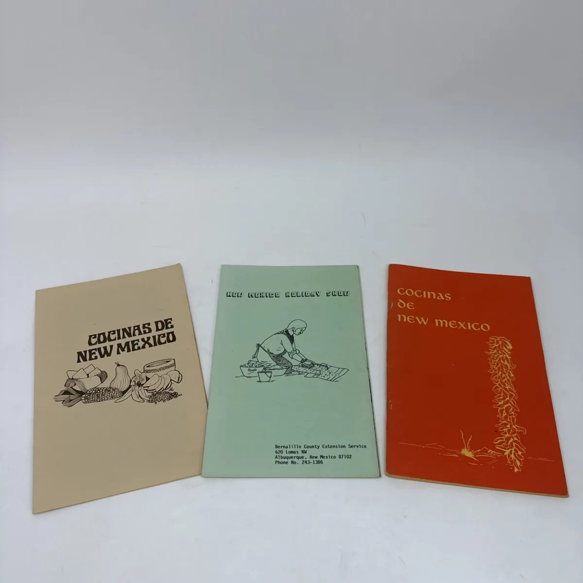 Three saddle-stitched soft-cover cookbooks lying side-by-side on white background: a tan Cocinas de New Mexico cover with stylized vegetables, a green Bernalillo County Extension Service New Mexico Holiday Show cookbook, and an orange Cocinas de New Mexico variant with a chile ristra illustration.